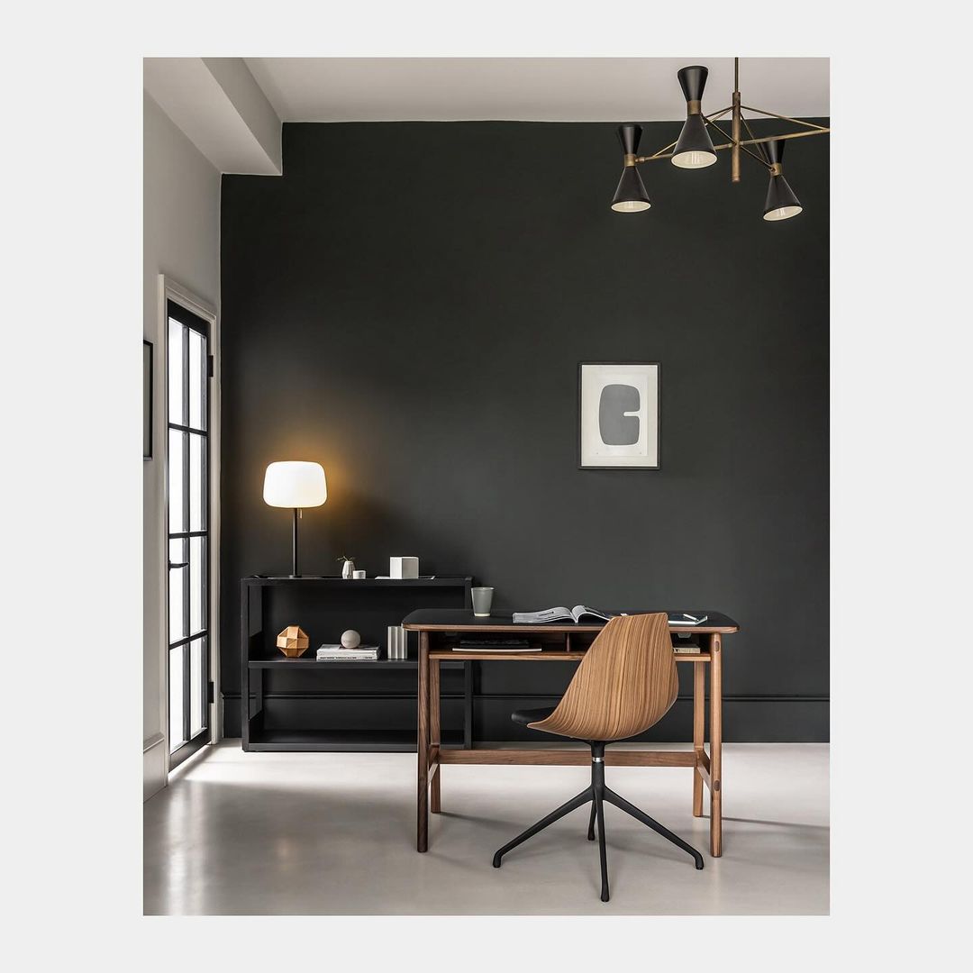 Interior of a home office with a wood desk and chair in front of a dark green wall, illuminated by natural light and a table lamp.