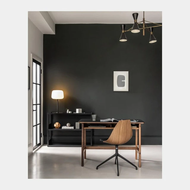 Interior of a home office with a wood desk and chair in front of a dark green wall, illuminated by natural light and a table lamp.