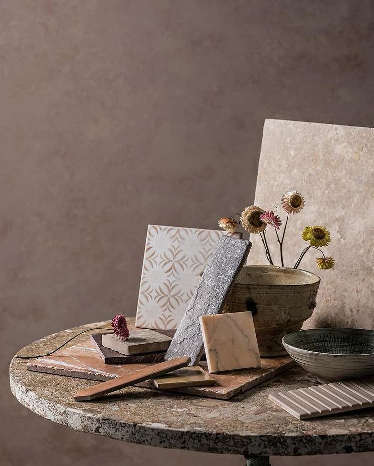 A collection of various tile samples arranged on a distressed round table, alongside a ceramic vase with dried flowers, set indoors.