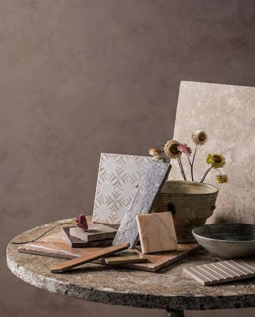 A collection of various tile samples arranged on a distressed round table, alongside a ceramic vase with dried flowers, set indoors.