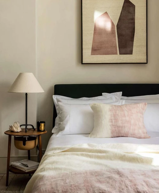 Bedroom interior featuring a made bed with white linens, a decorative pillow, and a patterned throw blanket. A wooden bedside table holds a lamp.