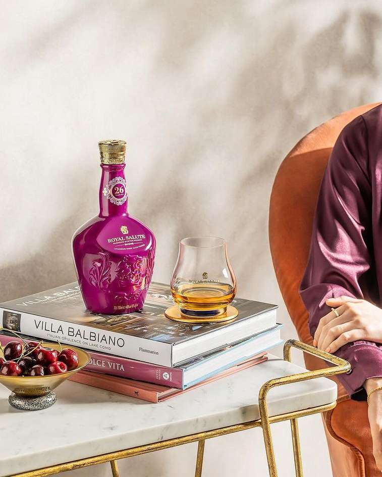 Royal Salute 26 year old whisky bottle and glass on books on a side table with cherries, and a person in robe sitting nearby.