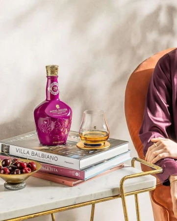Royal Salute 26 year old whisky bottle and glass on books on a side table with cherries, and a person in robe sitting nearby.