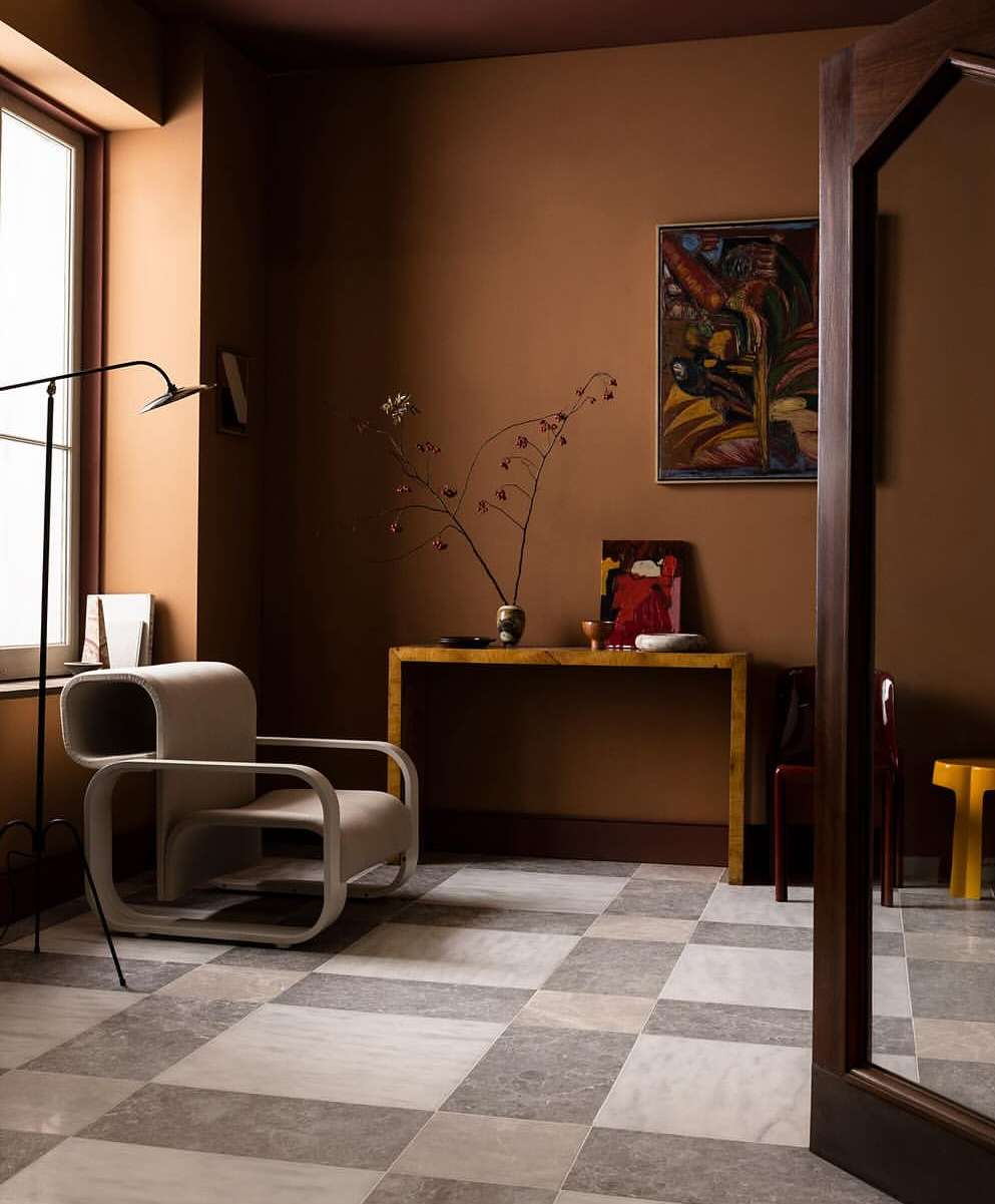 Interior view of a living space with a contemporary armchair, wooden console table, and checkered tile floor. Walls are painted in earth tones.