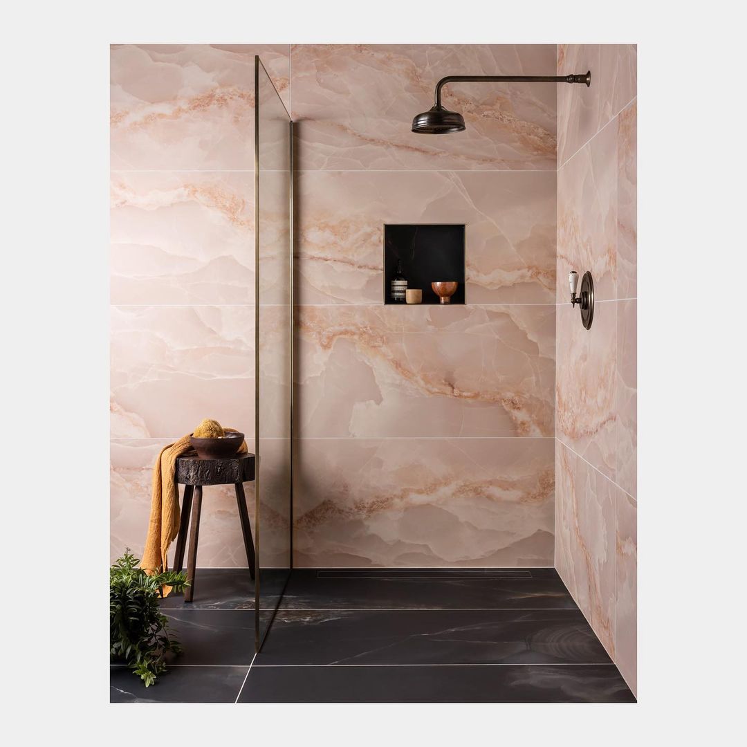 Bathroom interior featuring peach-colored marble tiles, black floor, showerhead, niche with toiletry items, stool with washcloth, and greenery.