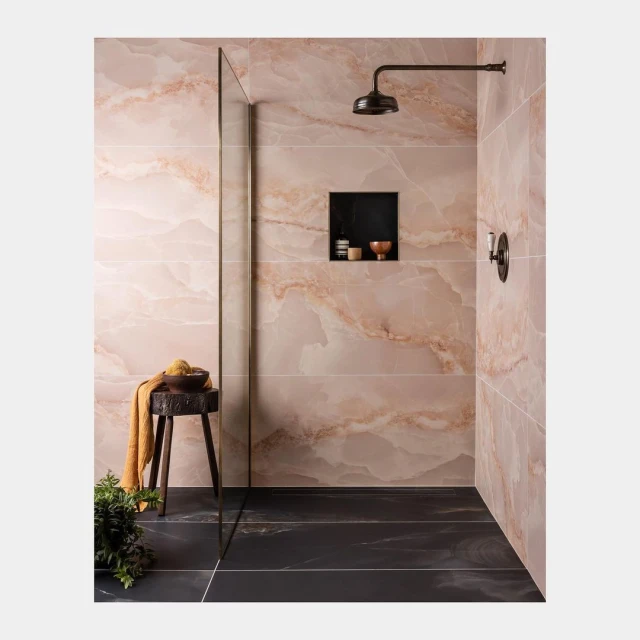 Bathroom interior featuring peach-colored marble tiles, black floor, showerhead, niche with toiletry items, stool with washcloth, and greenery.