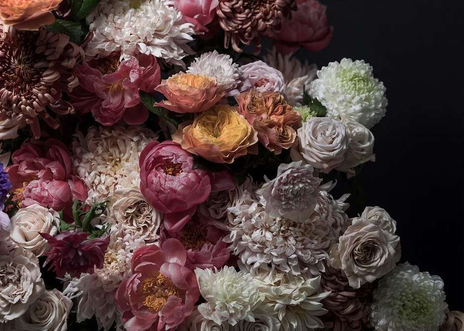 Close-up photograph of a lush floral arrangement with a variety of pink, peach, white, and brown chrysanthemums, peonies, and roses.