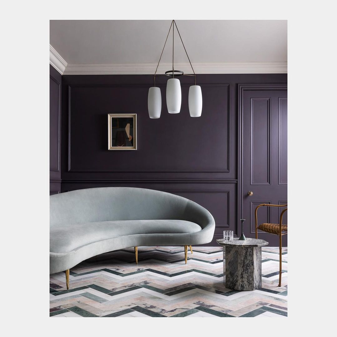 Contemporary living room interior: grey curved sofa, marble table, pendant lights, artwork, purple paneled wall, patterned floor.