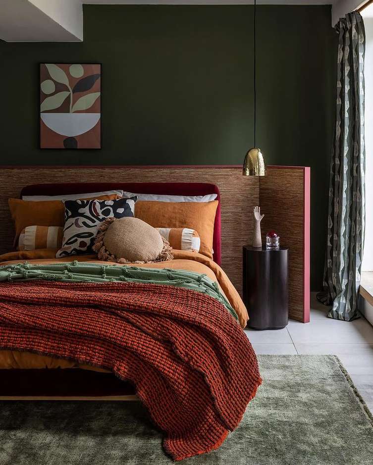 Bedroom interior featuring an orange bedspread, throw blanket, cushions, decorative pillows, dark green wall, and a patterned curtain.