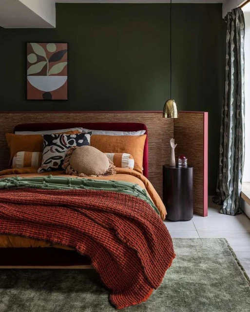 Bedroom interior featuring an orange bedspread, throw blanket, cushions, decorative pillows, dark green wall, and a patterned curtain.