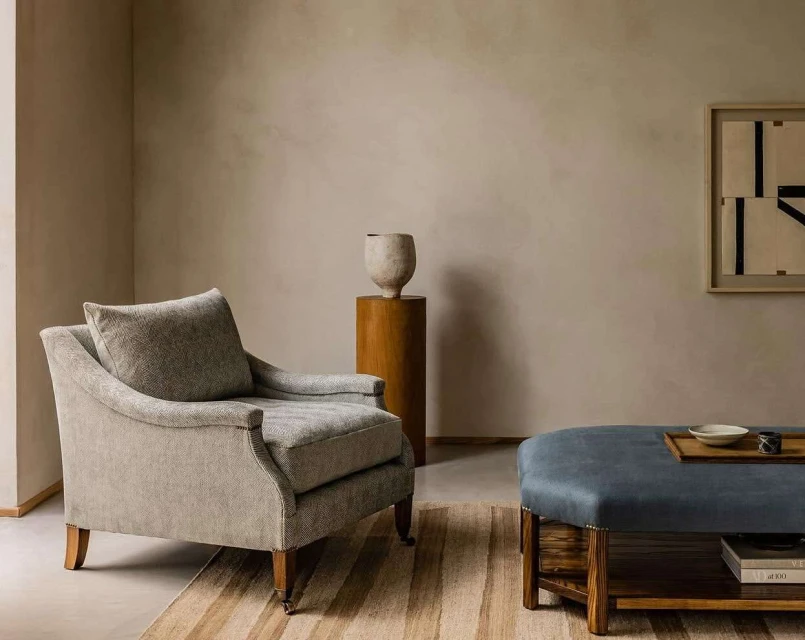 Interior design featuring a grey armchair, blue ottoman, ceramic vase on wooden pedestal, and framed graphic art piece in a neutral colour palette.