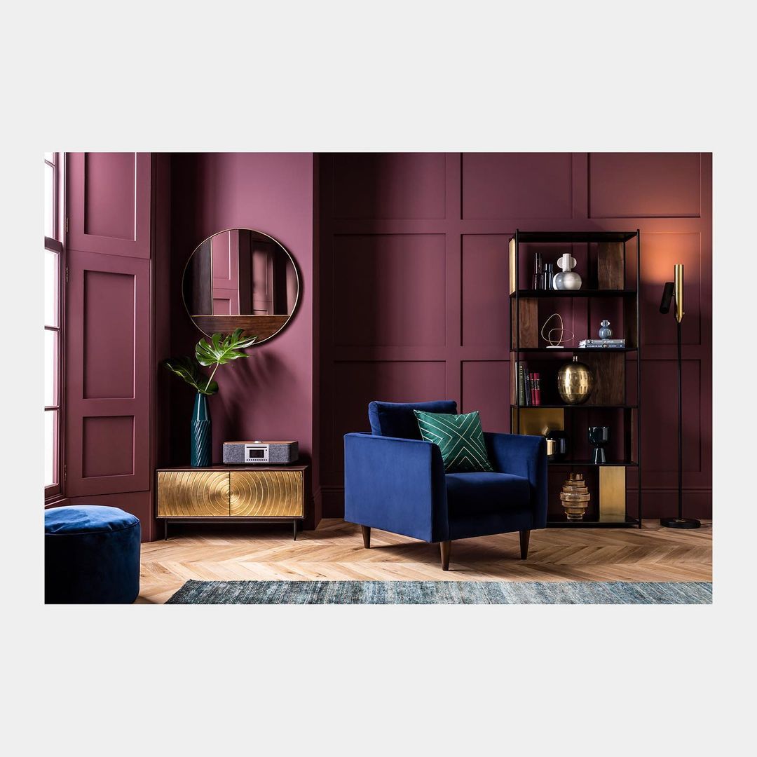 Interior design with a blue velvet armchair, gold cabinet, and dark red walls, complemented by a modern floor lamp and shelving unit.