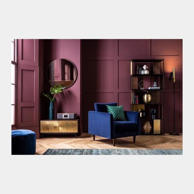 Interior design with a blue velvet armchair, gold cabinet, and dark red walls, complemented by a modern floor lamp and shelving unit.