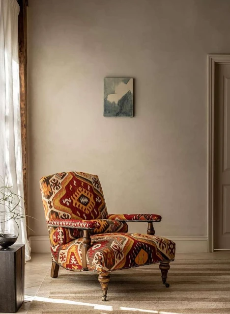 Vintage patterned armchair with wooden frame sits on a woven rug in a neutral-toned room with a door and abstract wall art.