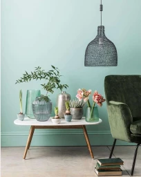 A white coffee table displaying glass vases and potted plants against a mint green wall with a velvet green armchair