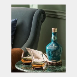 A teal ceramic whiskey bottle and two glasses of amber spirit on a green marble table with an armchair in the background.