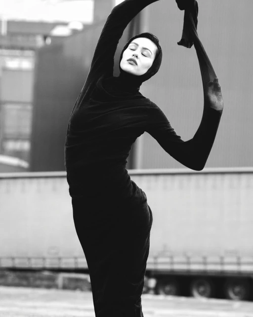 Artistic black-and-white portrait of a woman in a dark dress and head covering, captured outdoors against a backdrop of industrial architecture.