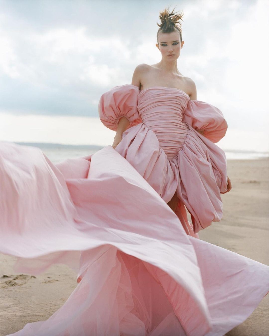 Fashion shot of a model in a pink, voluminous dress posing on a sandy beach with a cloudy sky backdrop, showcasing editorial style.