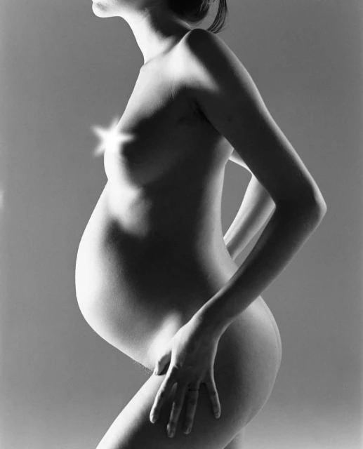 Monochrome studio shot of a nude pregnant woman in profile, showcasing her body and pregnancy against a soft grey backdrop.