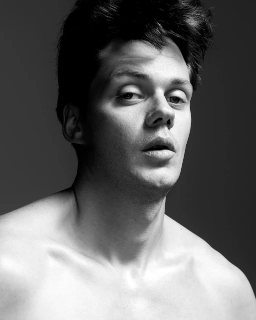 Black and white portrait of a shirtless adult male with dark hair against a grey background, showcasing his upper body and facial features.
