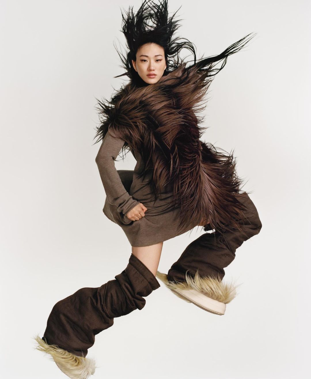 An Asian female fashion model strikes a dynamic pose in mid-air, wearing a brown dress, fur stole, and furry boots against a white background.