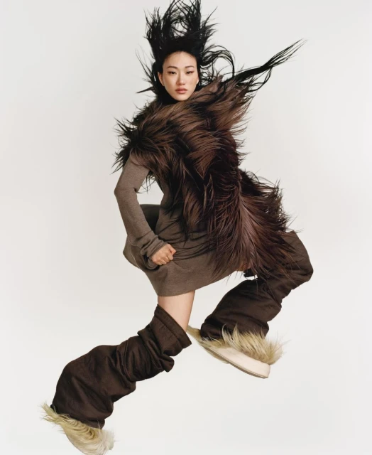 An Asian female fashion model strikes a dynamic pose in mid-air, wearing a brown dress, fur stole, and furry boots against a white background.