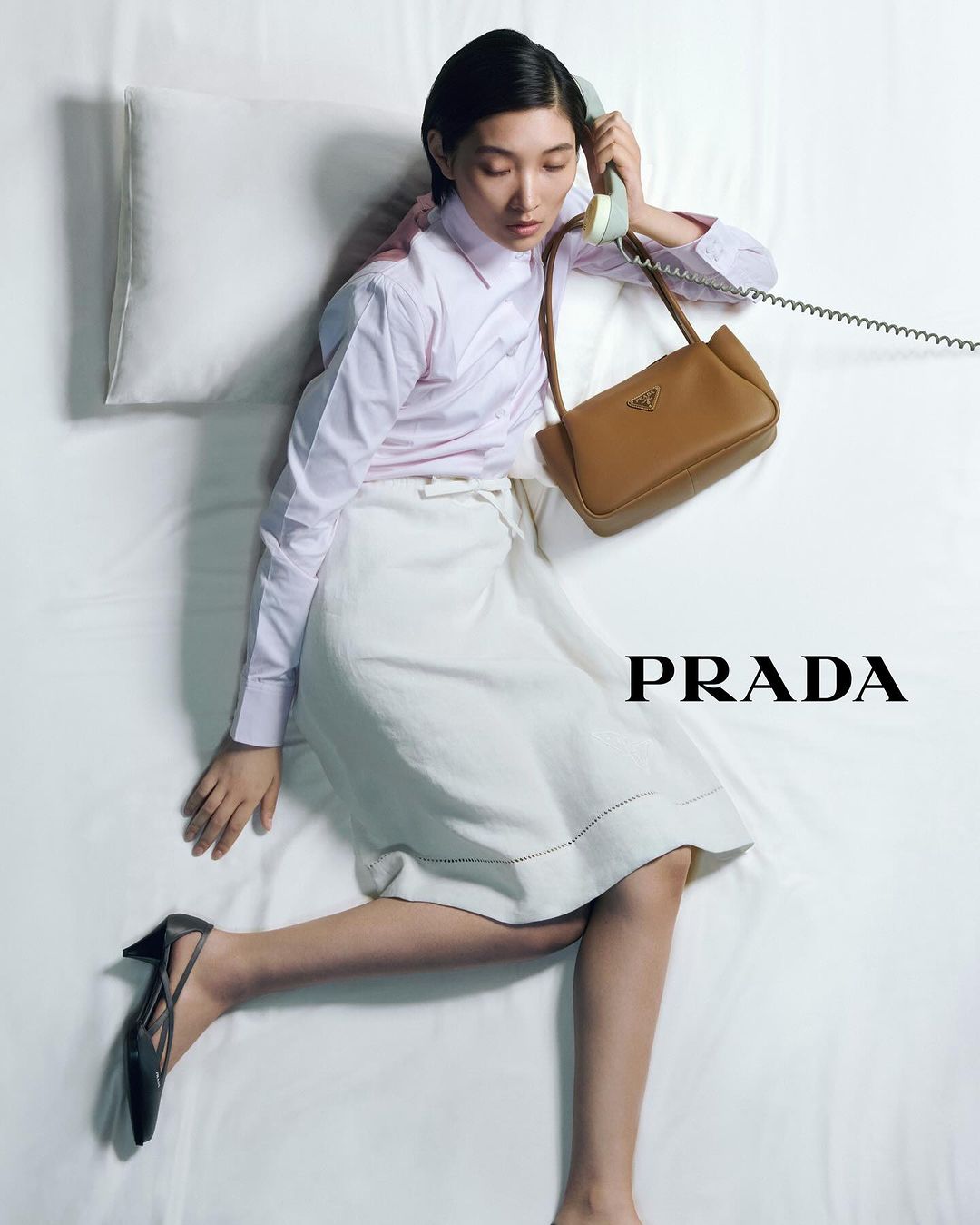 Woman lying on white bed, wearing pink shirt and white skirt, holding vintage telephone next to tan leather purse with Prada logo.