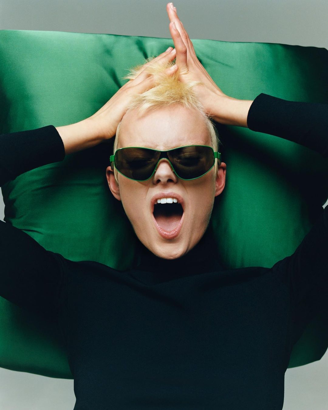 Expressive woman with hands on head, shouting, wearing sunglasses, turtleneck, and lying on a green cushion in a studio setting.