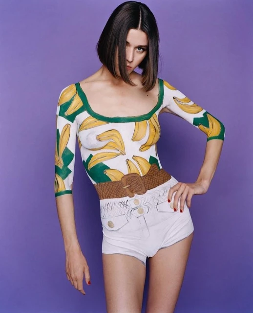 Brunette woman posing in a white bodysuit with banana pattern, wearing white denim shorts and a belt, set against a purple background.