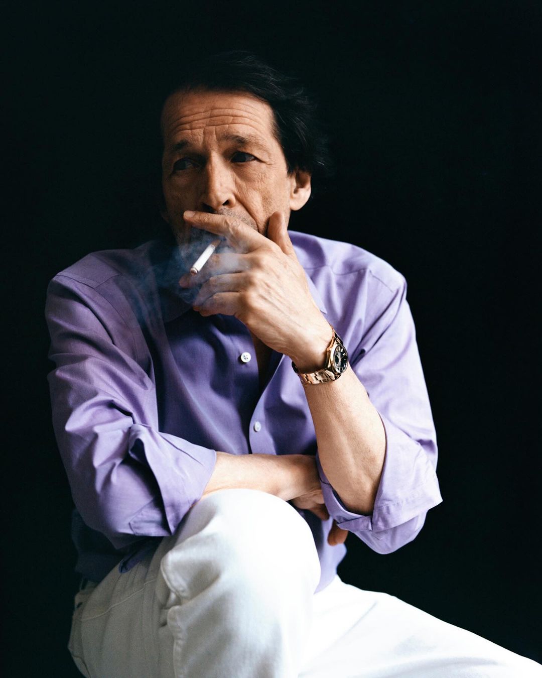 Portrait of a mature adult man smoking a cigarette while seated, wearing a button-down shirt, and white pants against a dark background.