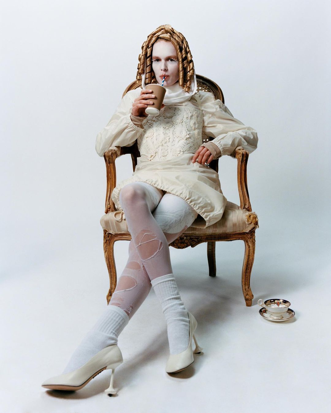 Model wearing an off-white dress and stockings, sits in an ornate chair while drinking from a paper cup with a straw in a studio.