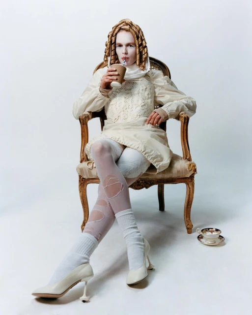 Model wearing an off-white dress and stockings, sits in an ornate chair while drinking from a paper cup with a straw in a studio.