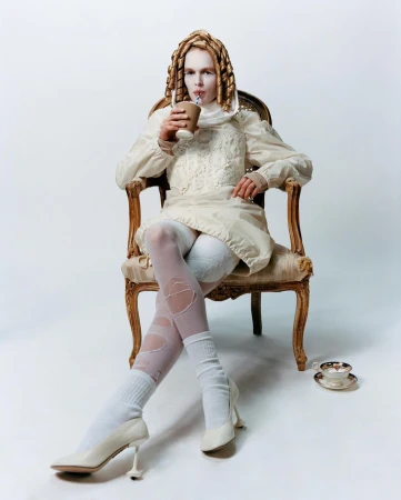 Model wearing an off-white dress and stockings, sits in an ornate chair while drinking from a paper cup with a straw in a studio.