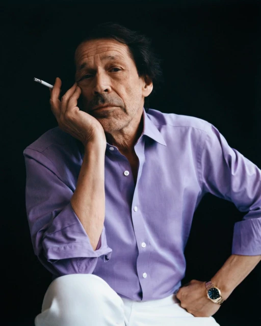 Portrait of an adult man with a cigarette, wearing a lavender shirt and white pants, sitting against a dark background in a studio.