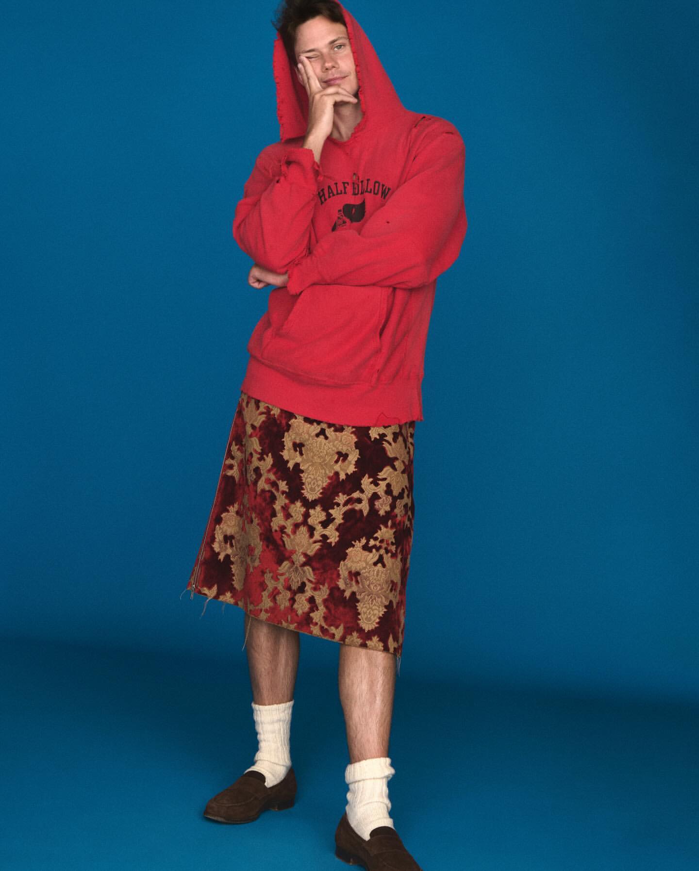 Young man with hoodie and floral skirt posing against blue background in studio lighting, looking off camera and holding chin.