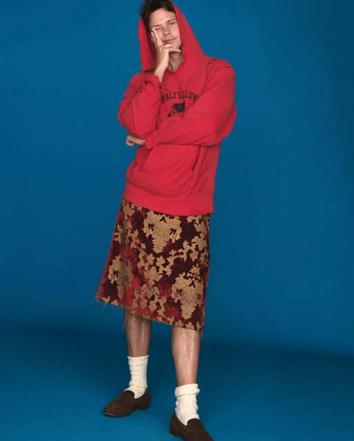 Young man with hoodie and floral skirt posing against blue background in studio lighting, looking off camera and holding chin.