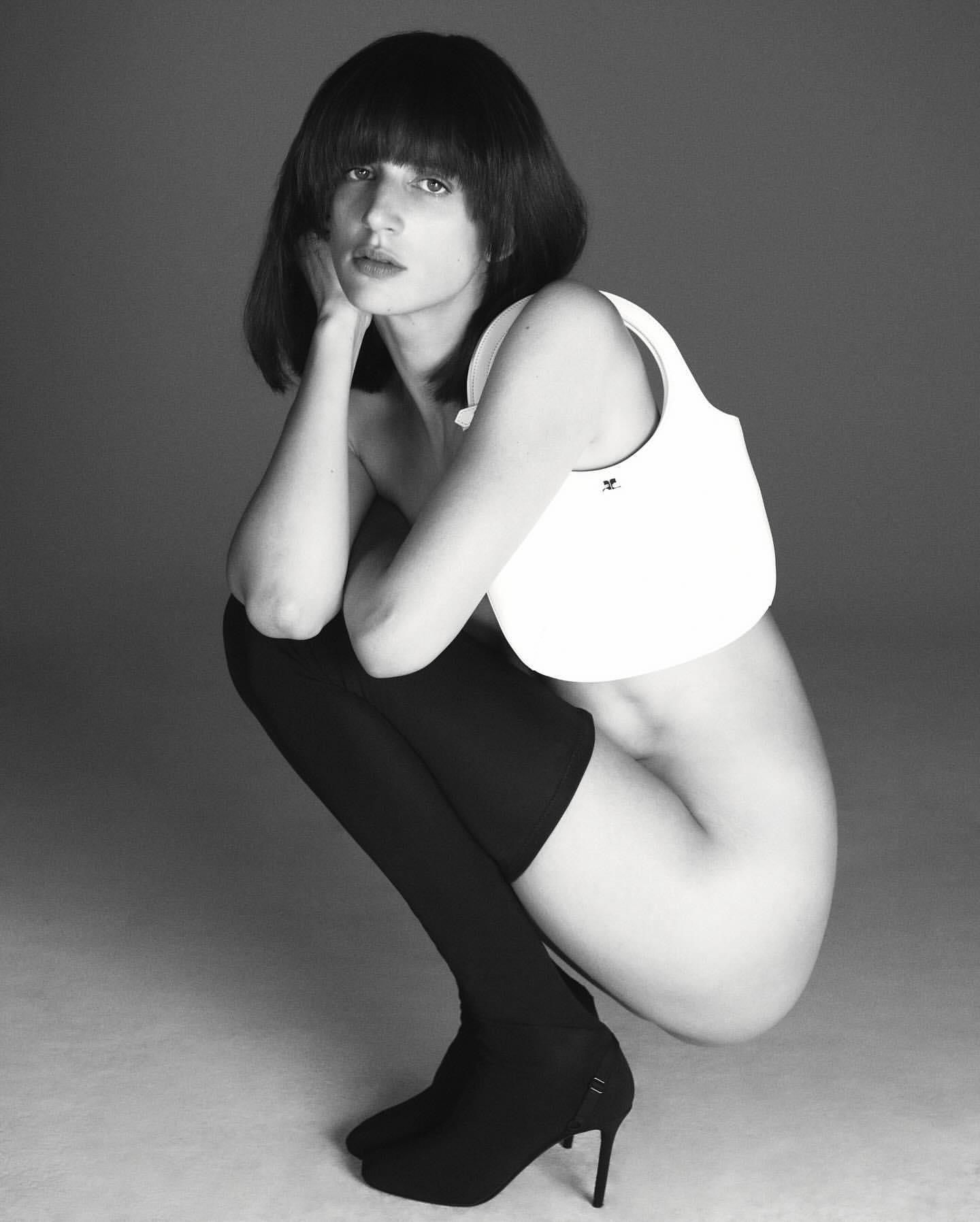 Black and white shot of a fair-skinned woman with black hair crouching, wearing thigh-high stockings, heels, and a small white bag.