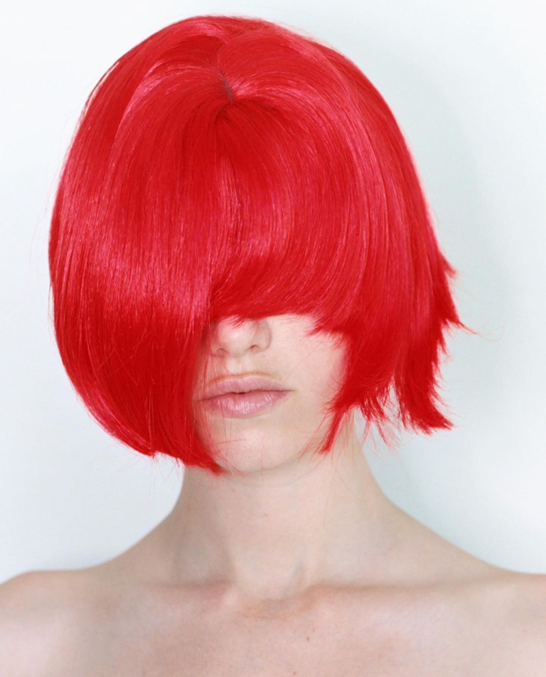 Close-up portrait of a model with a vivid red blunt bob wig, partly covering the face, against a plain white background.