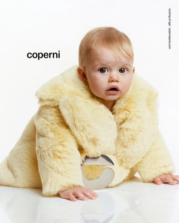Infant in a furry yellow coat crawls toward the camera, set against a bright white background, fashion brand name Coperni visible.