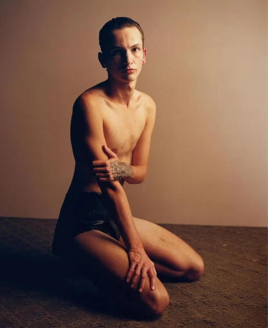 Nude human adult kneels indoors with neutral expression. Light skin, short hair. Arm tattoo visible. Soft, diffused studio lighting.