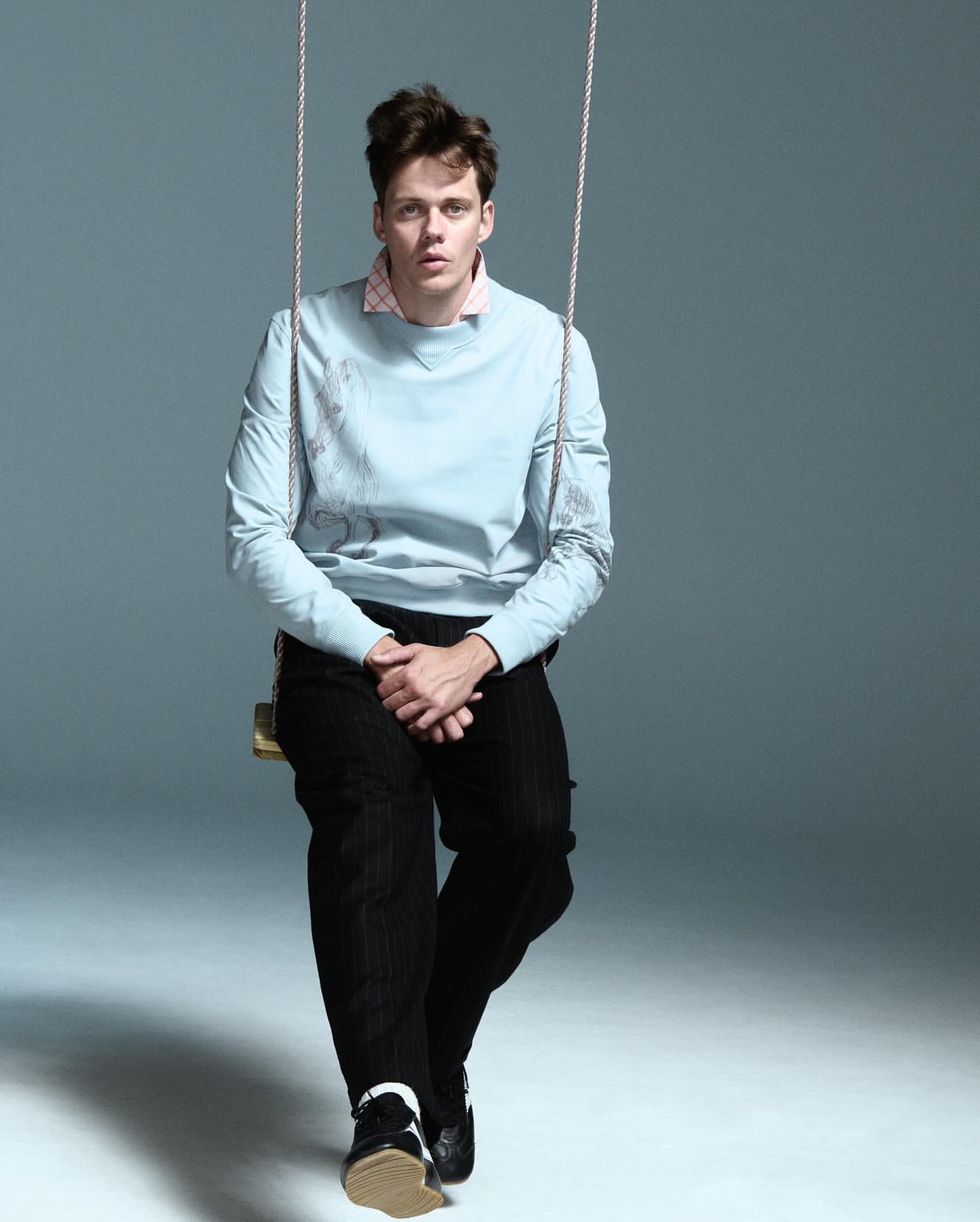 Man on a swing in a photo studio. He wears a blue sweatshirt, black pants, and black and white shoes. Neutral studio background.