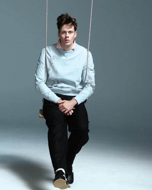 Man on a swing in a photo studio. He wears a blue sweatshirt, black pants, and black and white shoes. Neutral studio background.