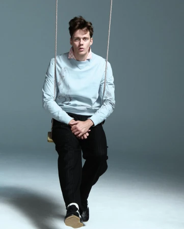 Man on a swing in a photo studio. He wears a blue sweatshirt, black pants, and black and white shoes. Neutral studio background.
