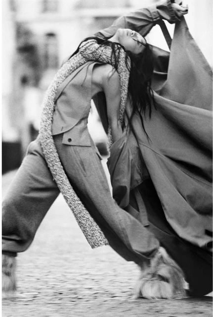 Monochrome fashion shot of a woman in a coat and scarf leaning backwards on a paved street with buildings in the background.