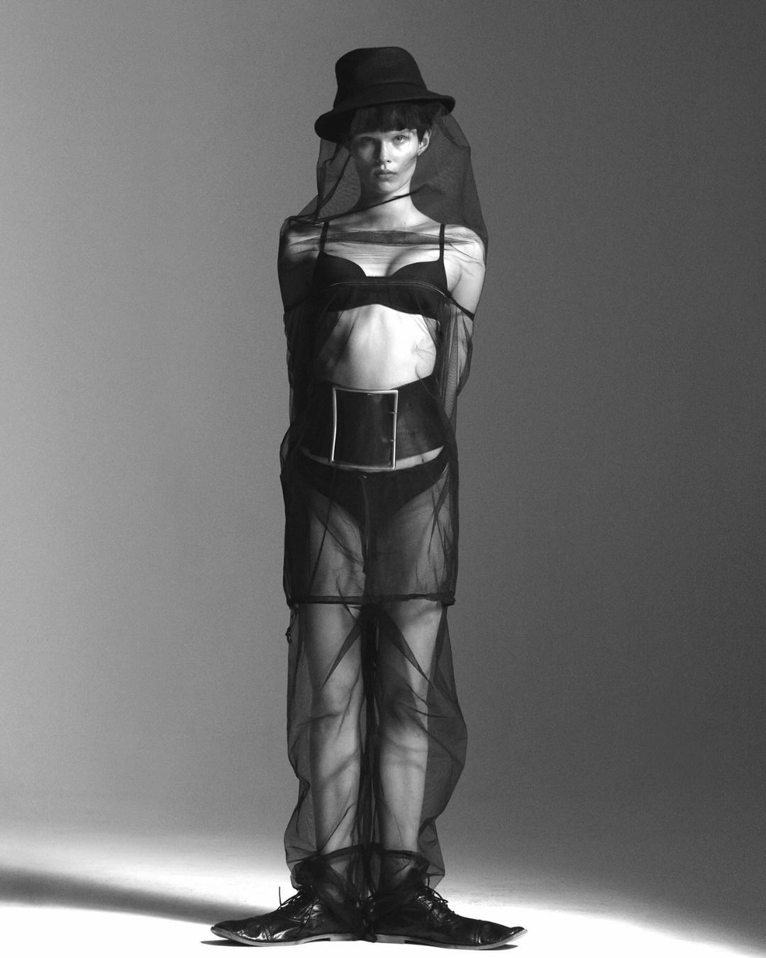 Monochrome fashion shot of a woman in lingerie and a hat, covered in black mesh, posing in a photo studio with soft, diffused lighting.