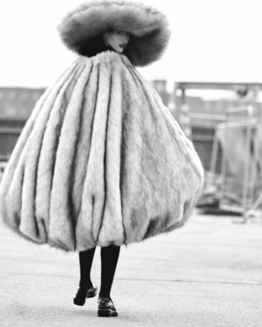 Monochrome fashion shot of a woman wearing oversized faux fur coat and hat, black tights, and chunky shoes walking on an empty road.