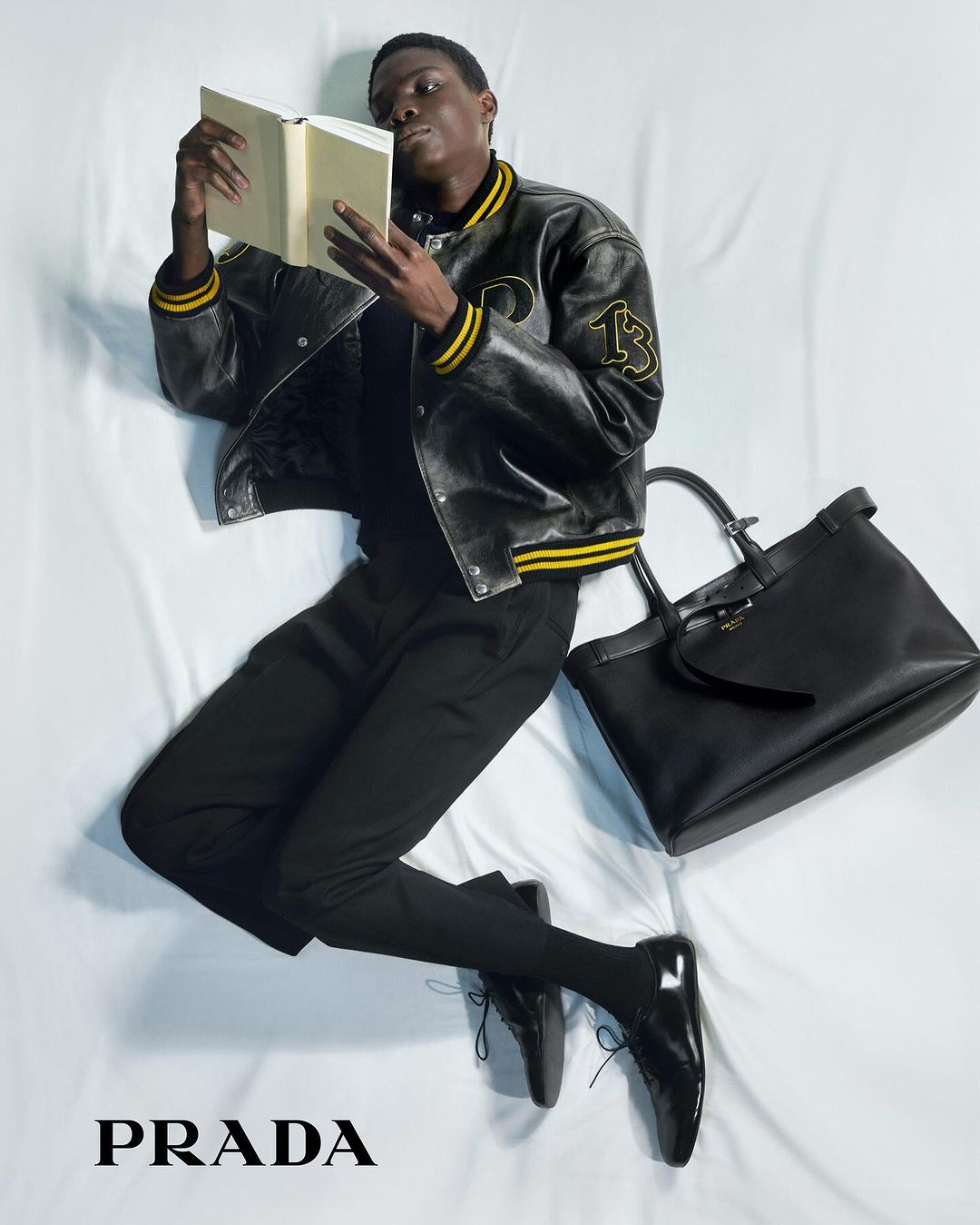 Fashion editorial featuring a Black male model in a varsity jacket lying on a white bed, reading a book next to a black leather handbag.