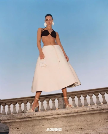 Model standing on a stone balustrade against a blue sky wearing a black top and white skirt holding a small white handbag