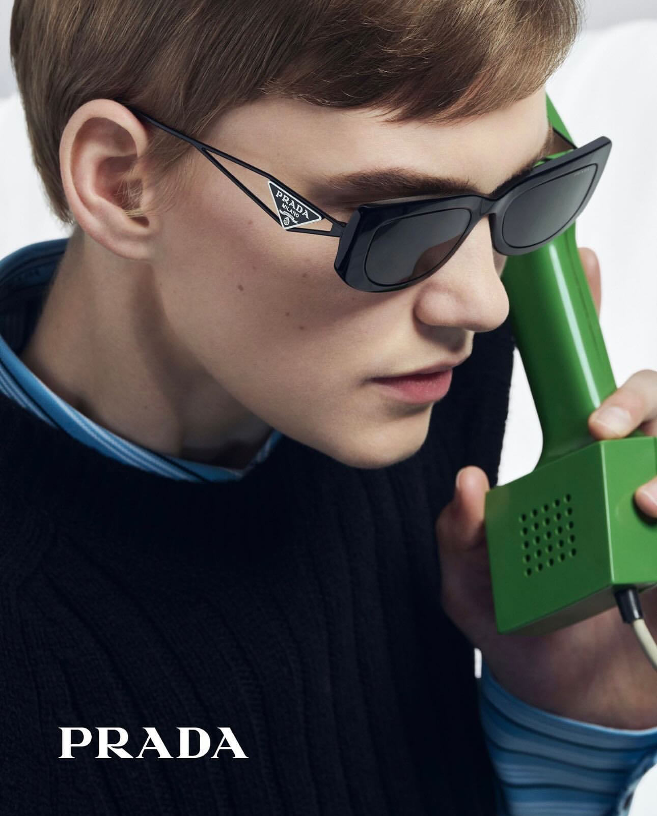 Close-up of a person wearing black Prada sunglasses and holding a green retro telephone receiver, showcasing a blend of vintage and modern.