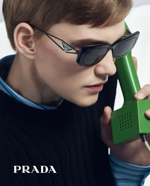 Close-up of a person wearing black Prada sunglasses and holding a green retro telephone receiver, showcasing a blend of vintage and modern.